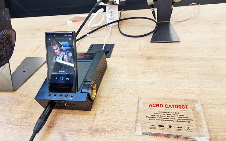 Astell & Kern ACRO CA1000T with an A&K player stood up in it. It's on a wooden table with some headphones partly visible on the left.