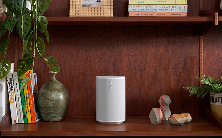 Sonos Era 100 on a shelf with books and a vase beside it and books on a shelf above it. The Era 100 is white.
