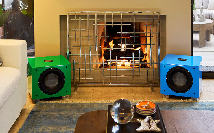The REL T9x special edition in green on the left and the blue on the right.  they are in front of a fireplace with a sofa to the left and objects on a coffee table in the foreground.