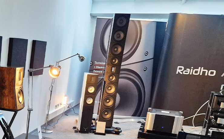 the Raidho TD6 floorstanding loudspeaker beside a white Raidho X2.6 loudspeaker with a Moon power amp on a low stand the other side. there are lamps and a Raidho backdrop in the photo too along with a bookshelf speaker on a speaker stand. The photo was taken at the Munich 2024 HiFi Show
