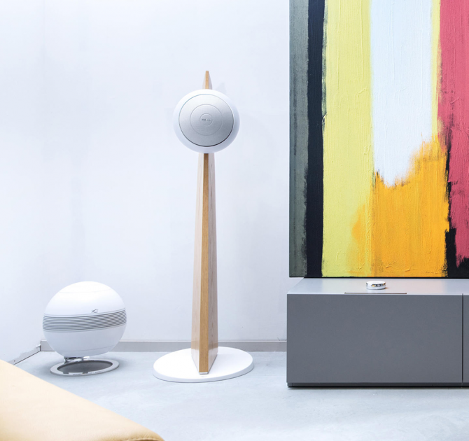Cabasse Baltic 5 Sub System (on stand).  The sub is white.  The Baltic 5 is white on a wooden stand.  It's beside a low sideboard with abstract art on the wall.