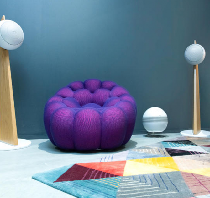 Cabasse Baltic 5 Sub System (on stand).  The sub is white.  the Baltic 5s are white with wooden stands.  There's a geometric rug on the floor.  The wall is painted dark grey and there's a purple chair that looks like a solid beanbag.