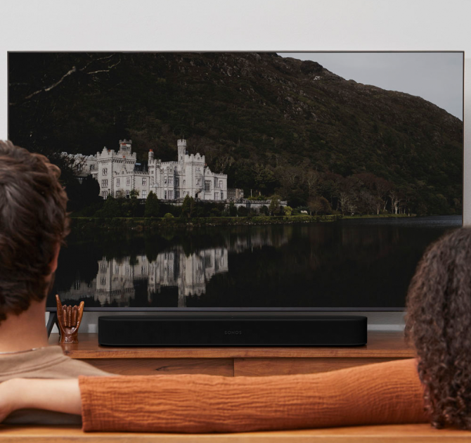 SONOS Beam (Gen 2) in black.  there's a man in the foreground on the left and a woman on the right.  She has her arm on the back of the sofa.  the tv is showing a picture of a castle reflected in a lake with a hill behind it.