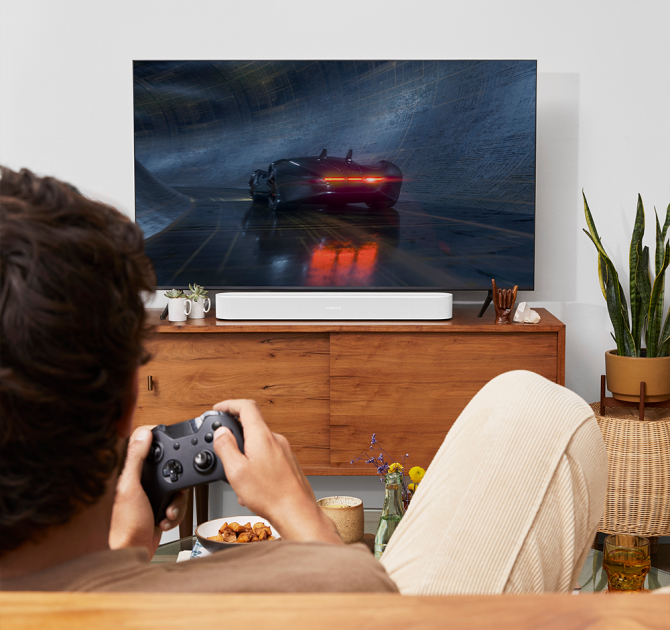 SONOS Beam (Gen 2) in white on top of a tv cabinet.  there's a man in the foreground holding a gaming controller.  He's playing a driving game.  there's a mother-in-law's tongue plant to the right of the tv cabinet.
