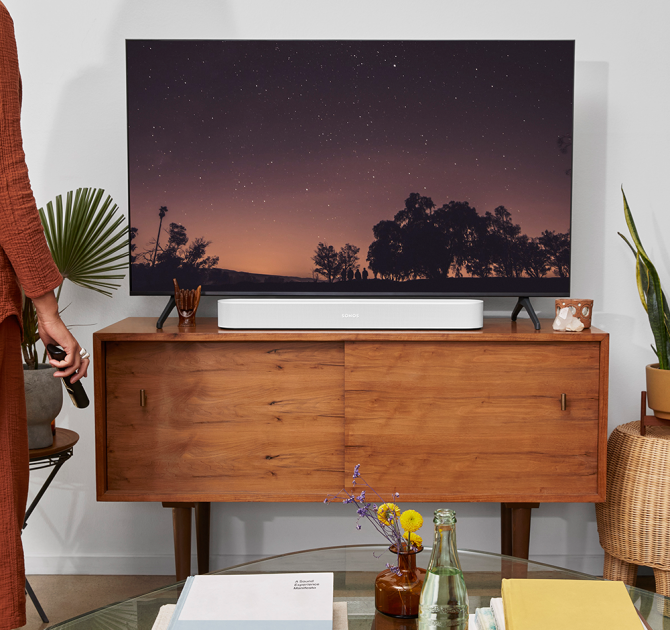 SONOS Beam (Gen 2) in white on top of a wooden tv unit.  there's a woman standing with the tv remote in her hand.  She's on the right of the image and only part of her can be seen.  the tv is showing something that involves a sunset with a line of trees in silhouette.