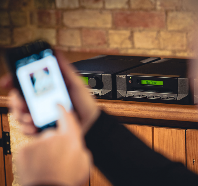 Cyrus CD T CD Transport on a wooden unit with a phone in the foreground being held and controlling the music.