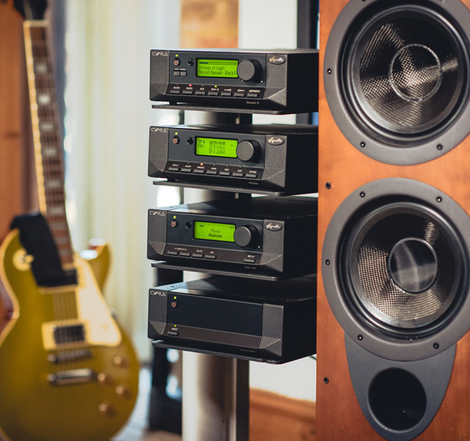 Cyrus Stereo 200 Power Amplifier on a stand with other pieces of Cyrus equipment with a speaker to the right and a guitar to the left.