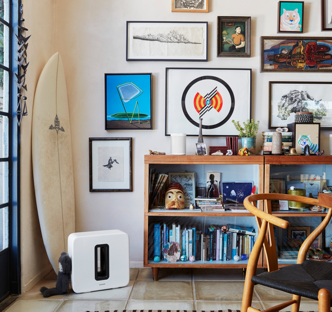 SONOS Sub in white on the floor in front of a surf board, a wall with lots of framed pictures and a cluttered shelving unit.