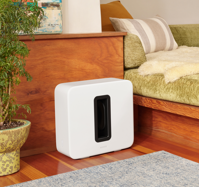 SONOS Sub in white, on the floor between a tall houseplant and a sofa