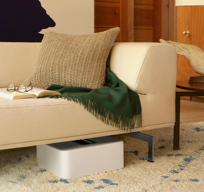 SONOS Sub in white laying down under a sofa that has an open book and a pair of glasses laying on it.