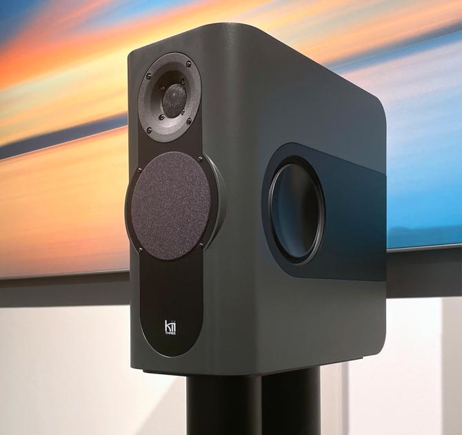 A dark grey rectangular shaped speaker on a stand with a colourful abstract print behind it.