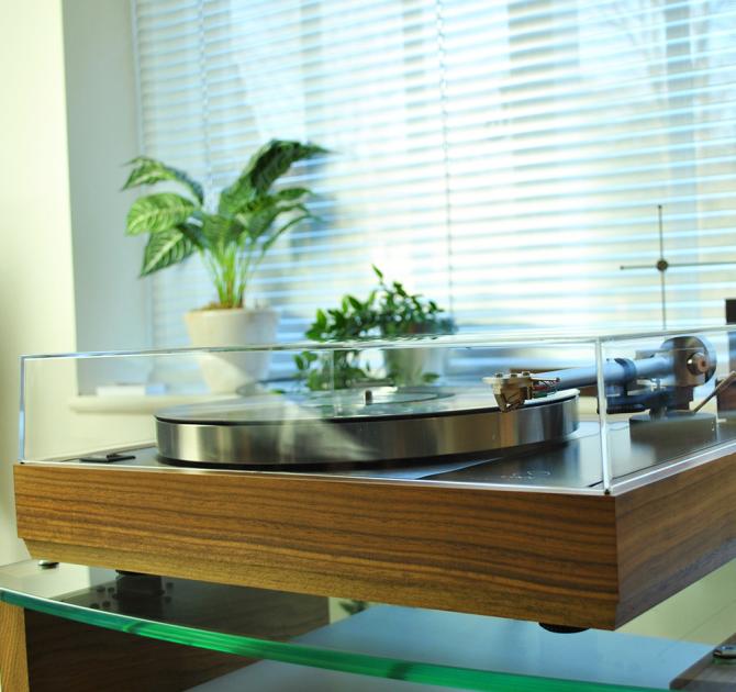 Record player with the lid on.  It's on a glass shelf in front of a window.  The windowsill has two plants on it that are out of focus.  The focus is on the turntable.