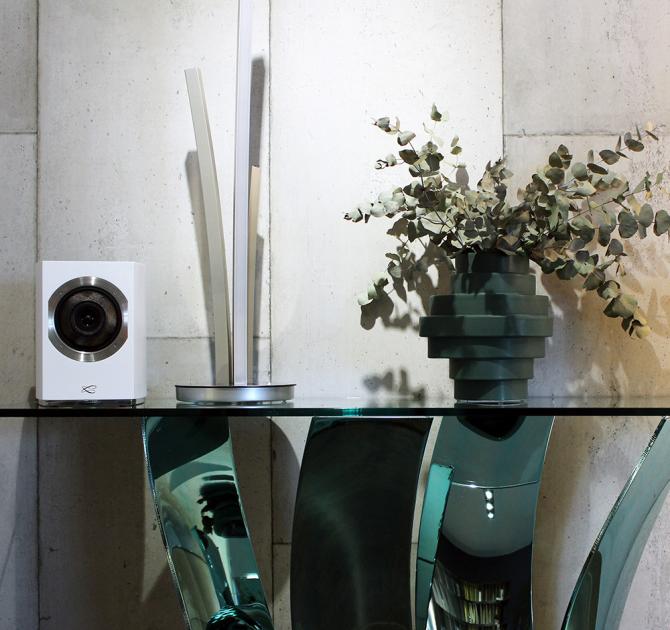 A Cabasse Rialto Loudspeaker in white on top of a glass table with a vase of eucalyptus on the right.