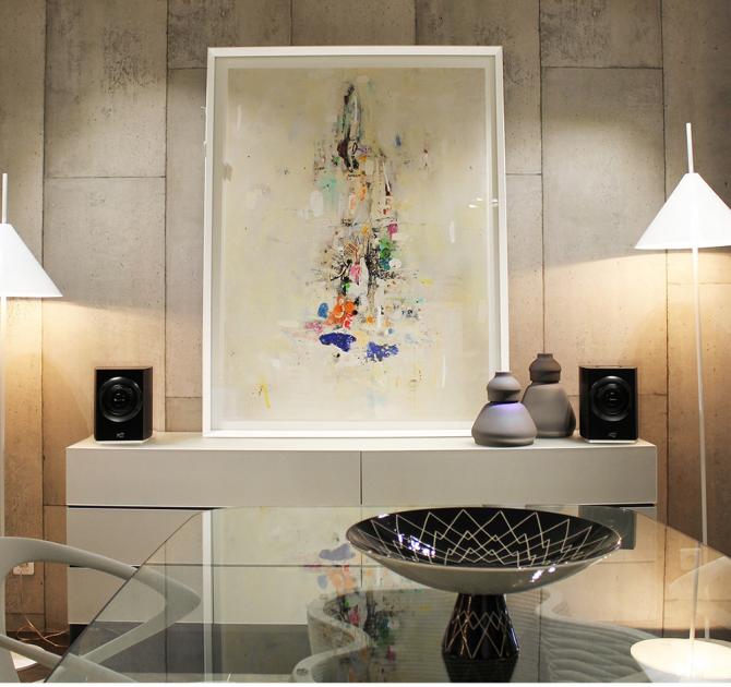 A pair of Cabasse Rialto Loudspeakers in black on top of a unit either side of a piece of abstract art with some vases.