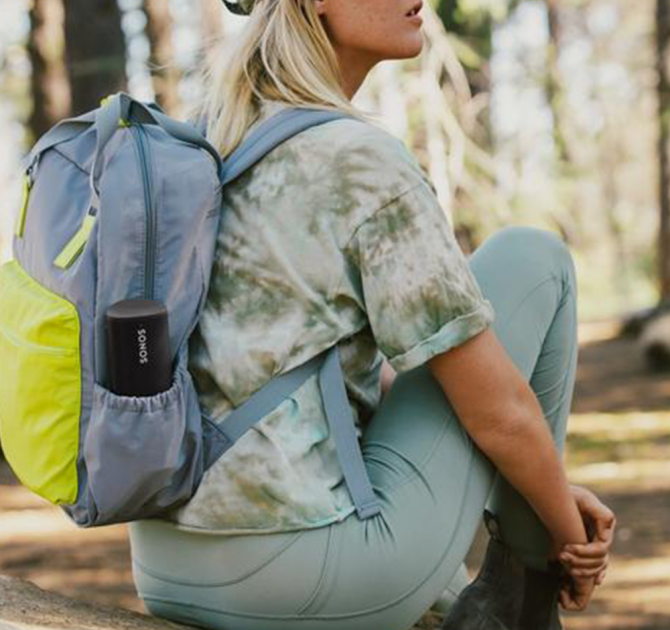 SONOS Roam SL in black in the side pocket of a backpack.  The woman is sitting on the floor.