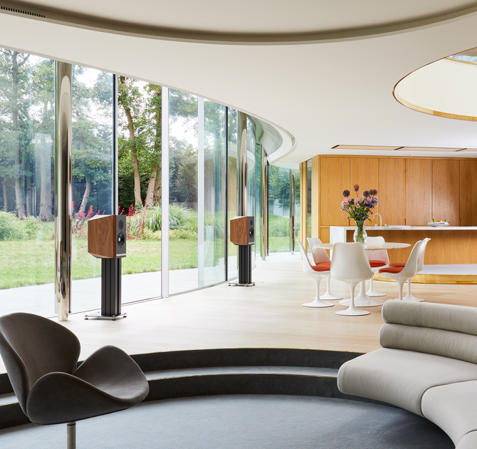 A pair of Titan 505 Speakers on stands in a large, modern living area.  They are in front of large glass doors opening out onto a garden.