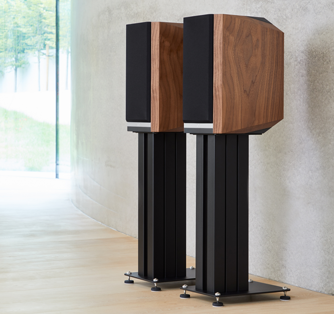 A pair of Titan 505 speakers on stands with grills on in front of a polished concrete wall.