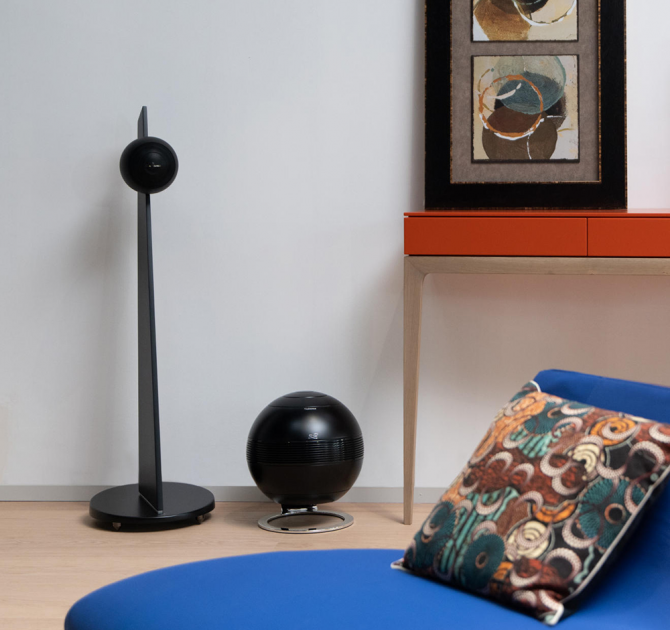 Cabasse iO3 Loudspeaker on stand in black with a Pearl Sub in black beside it.  There's a table beside it and a picture resting on the table.  In the foreground is a blue chair with a colourful cushion.