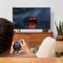 SONOS Beam (Gen 2) in white on top of a tv cabinet.  there's a man in the foreground holding a gaming controller.  He's playing a driving game.  there's a mother-in-law's tongue plant to the right of the tv cabinet.