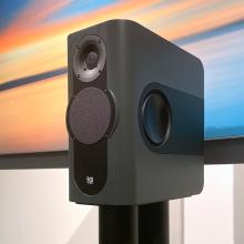 A dark grey rectangular shaped speaker on a stand with a colourful abstract print behind it.