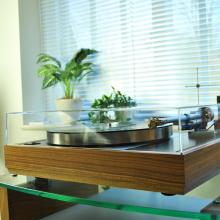 Record player with the lid on.  It's on a glass shelf in front of a window.  The windowsill has two plants on it that are out of focus.  The focus is on the turntable.