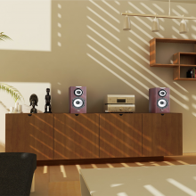 A pair of Cabasse Minorca Loudspeakers with a hifi between on a sideboard with black statues.  There's a large painting on the wall and wooden shelves on the wall.  There's a table in the foreground with two black sofas