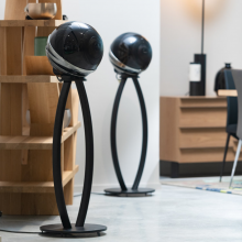 A pair of Cabasse Pearl Loudspeaker in black on stands in a living area with shelves and a chest of drawers