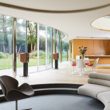 A pair of Titan 505 Speakers on stands in a large, modern living area.  They are in front of large glass doors opening out onto a garden.