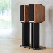 A pair of Titan 505 speakers on stands with grills on in front of a polished concrete wall.
