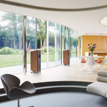 A pair of Kudos Titan 707 speakers with grills off in a large, modern living area in front of a wall of glass doors with a garden beyond.