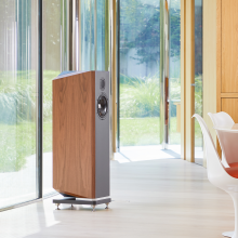A single Kudos Titan 707 speaker with grills off in a large, modern living area in front of a wall of glass doors with a garden beyond.