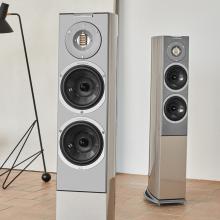A pair of tall, slim speakers on a wooden floor with a lamp to the left of the image - a tall angled style lamp